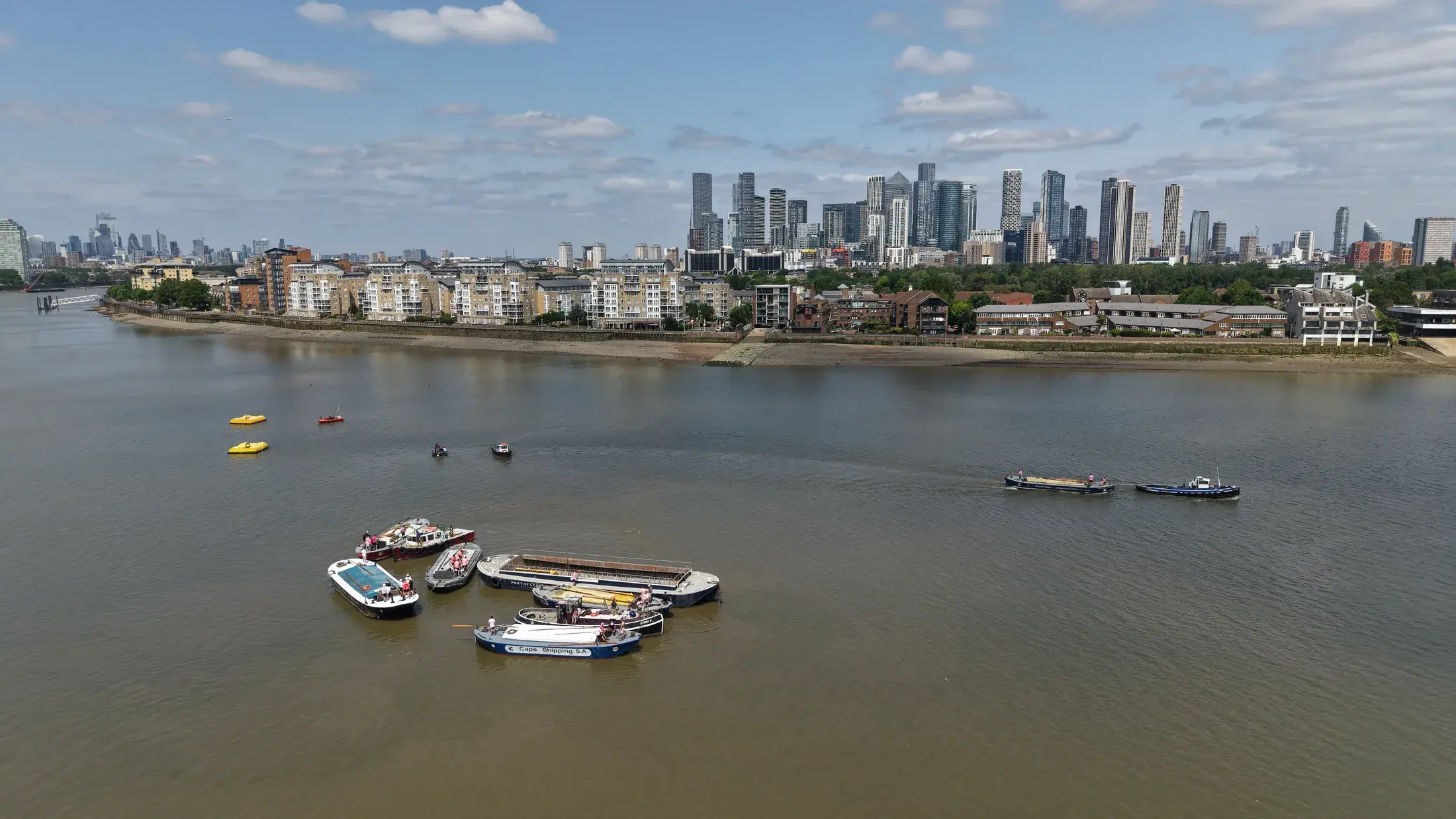 Aerial view over Greenwich Reach of Thames barges and tugs gathering before the 2026 Historic Barge Driving Race, with the Canary Wharf skyline beyond.