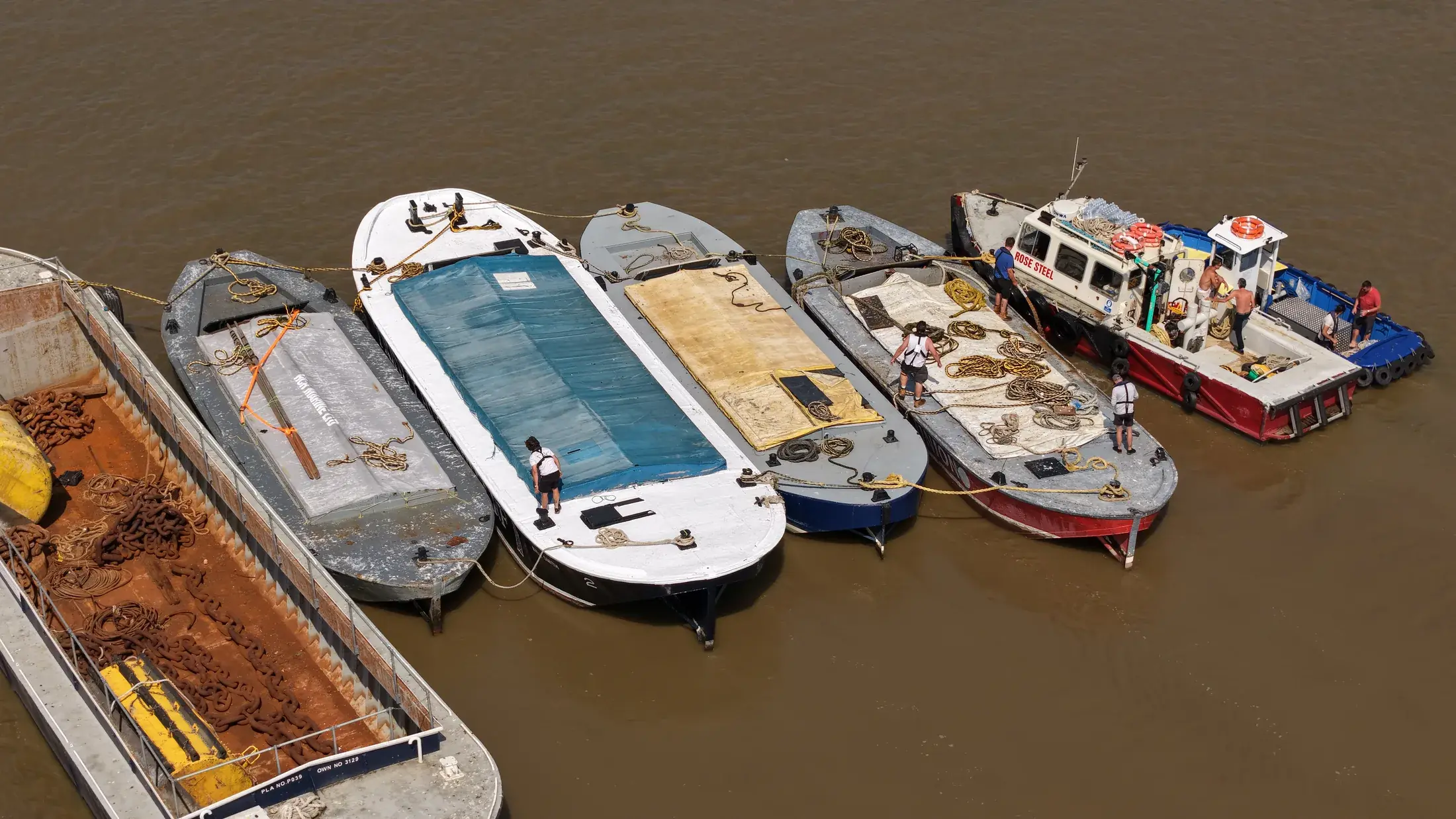 Aerial photograph looking down on Thames barges rafted alongside the tug Rose Steel, with crew members coiling ropes on deck.