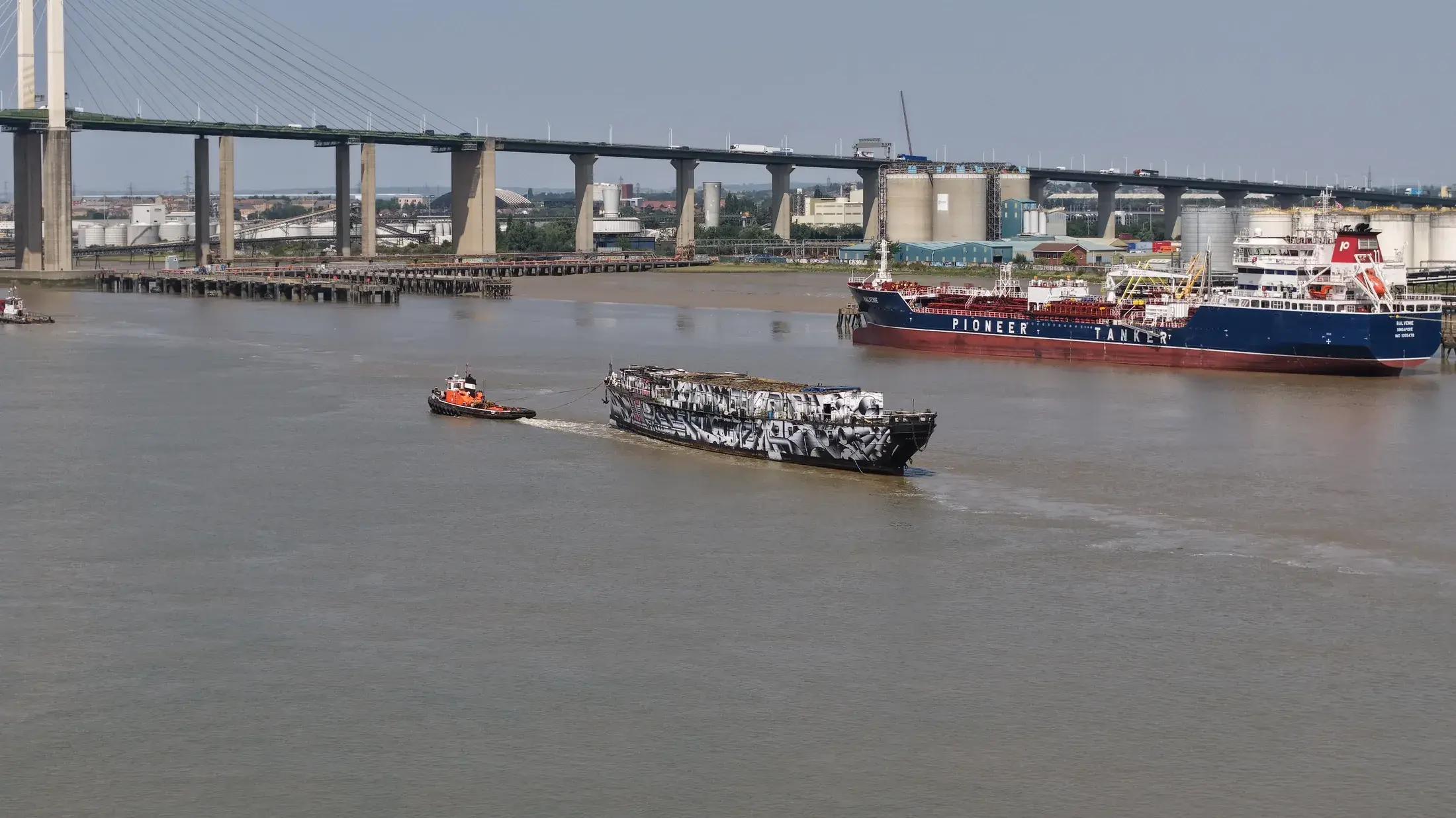 Aerial photograph of HMS President 1918 under tow on the Thames, passing a moored tanker at a riverside jetty near Dartford.