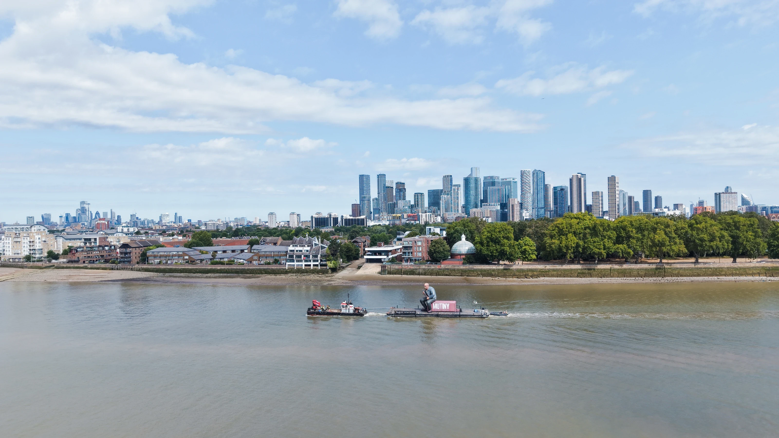 The tug Emilia D and the Mutiny container pontoon on the River Thames with the Canary Wharf skyline in the distance