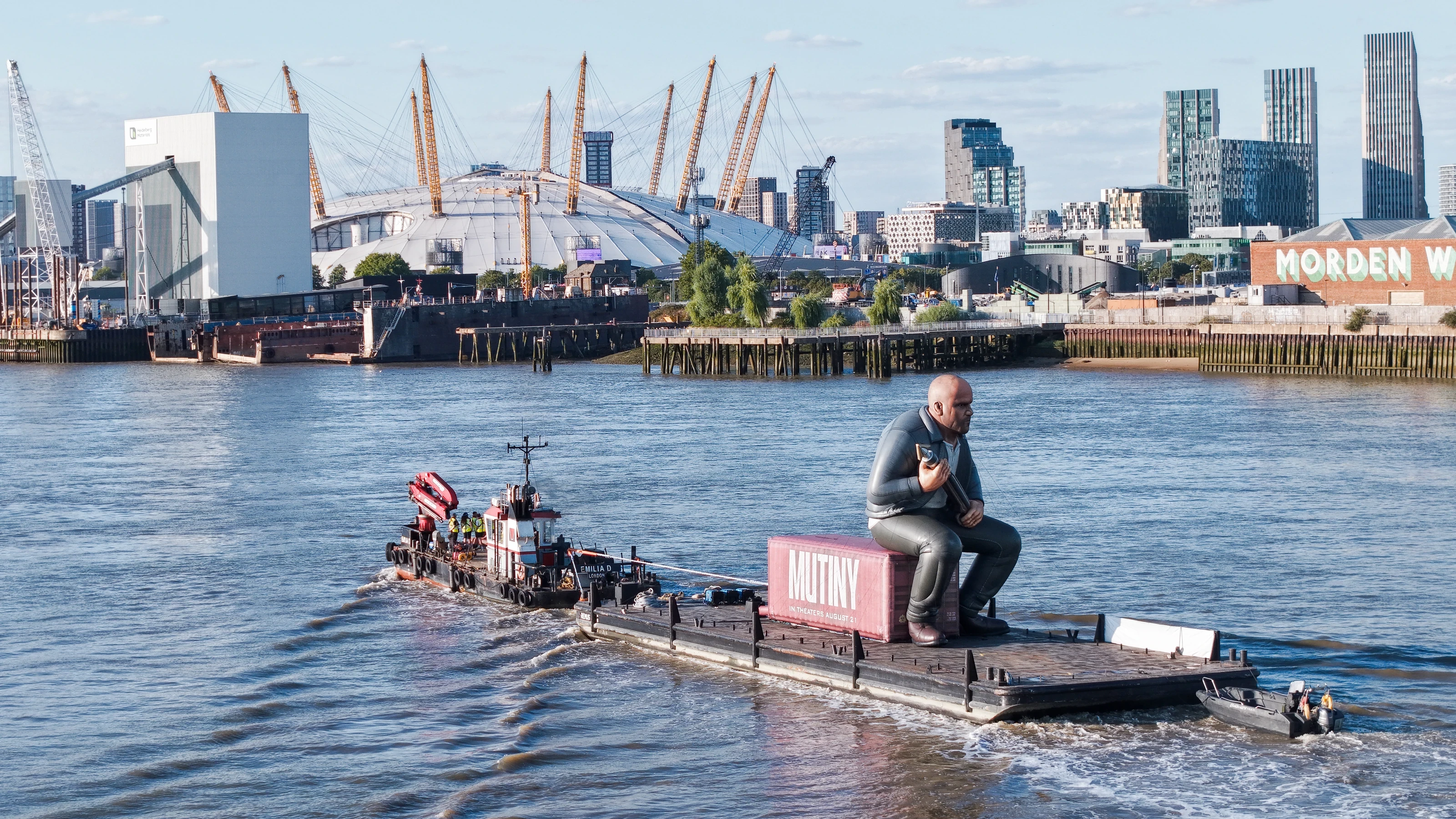 Aerial view of the inflatable Jason Statham seated on the pink Mutiny shipping container aboard a flat-top pontoon under tow on the River Thames