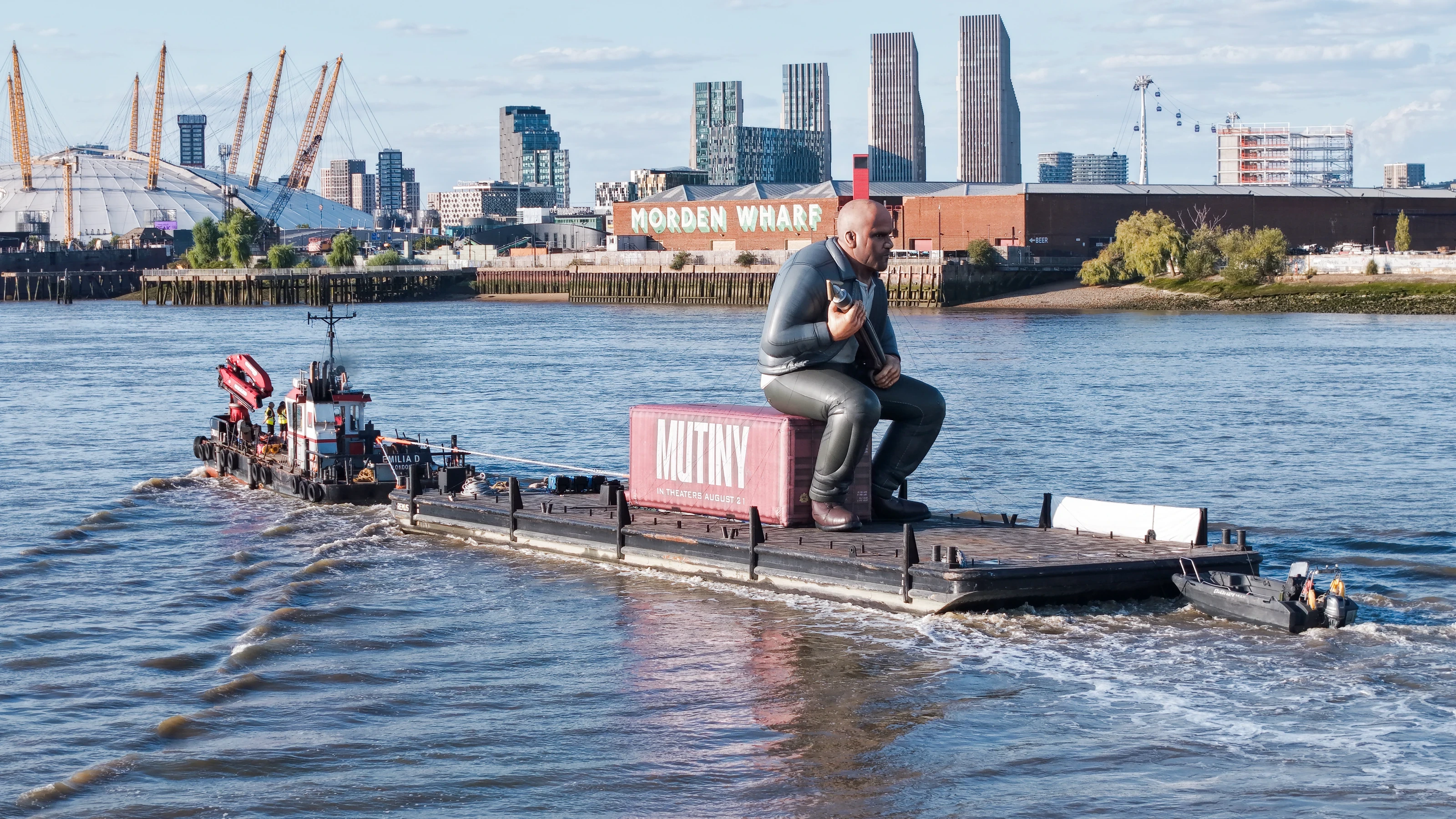 Evening aerial photograph of a giant inflatable Jason Statham seated on a pink Mutiny shipping container aboard a pontoon under tow past the Greenwich Peninsula, with the O2 arena behind.