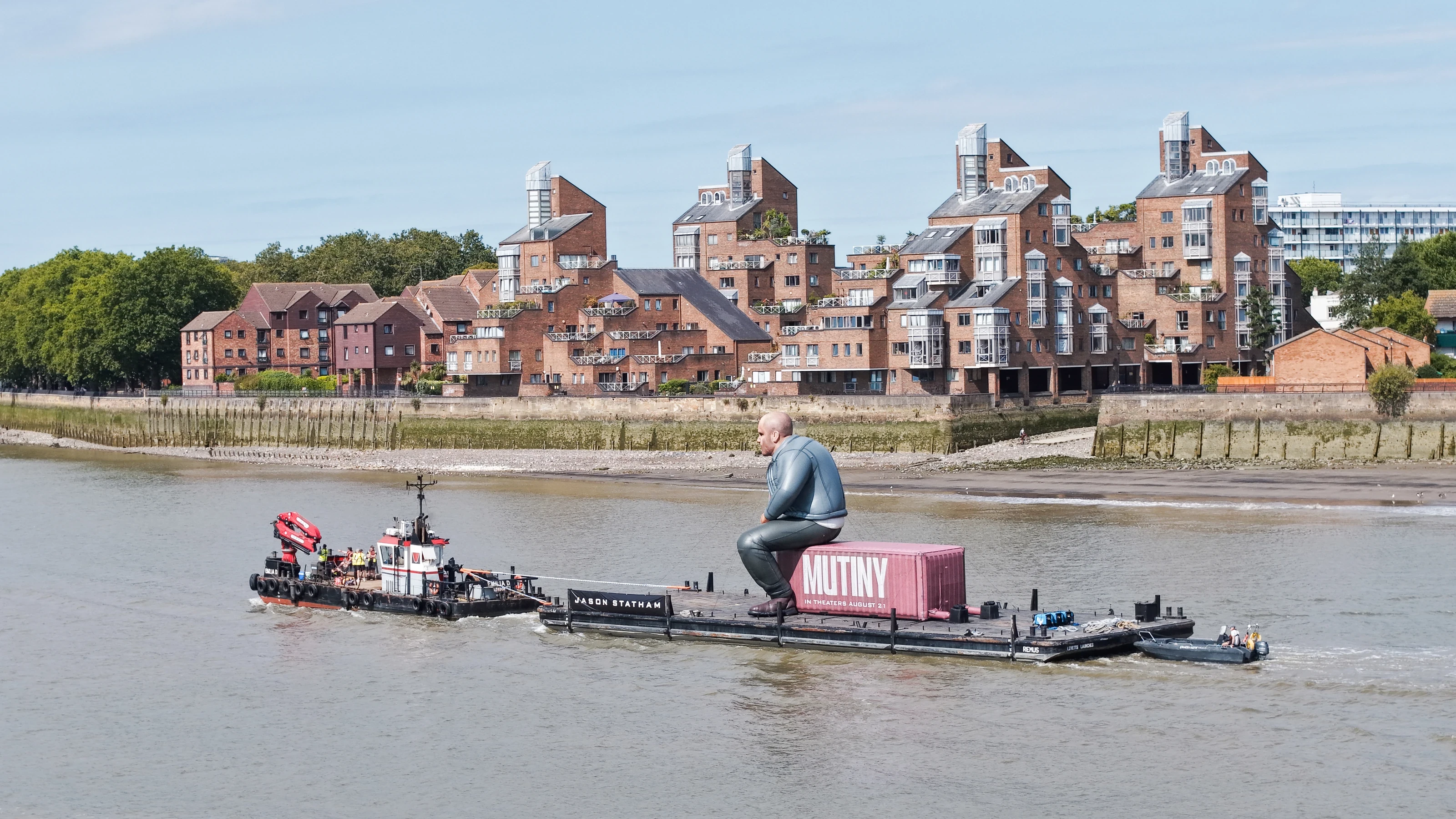 The multicat Emilia D towing a pontoon carrying a giant inflatable Jason Statham seated on a pink Mutiny shipping container on the River Thames in London.