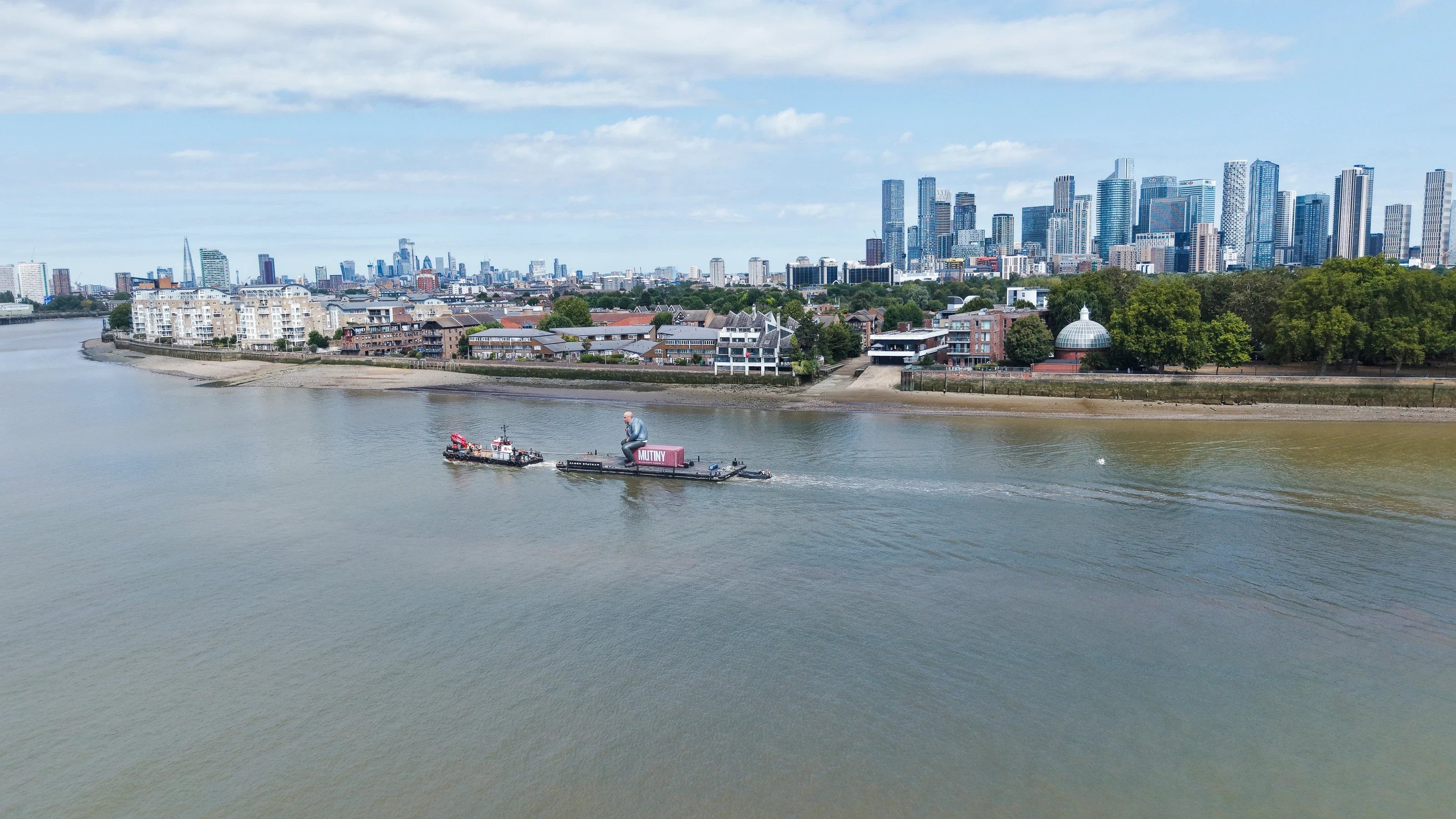 Wide aerial photograph of the Mutiny activation tow in Greenwich Reach, with the Isle of Dogs and the City of London skyline behind