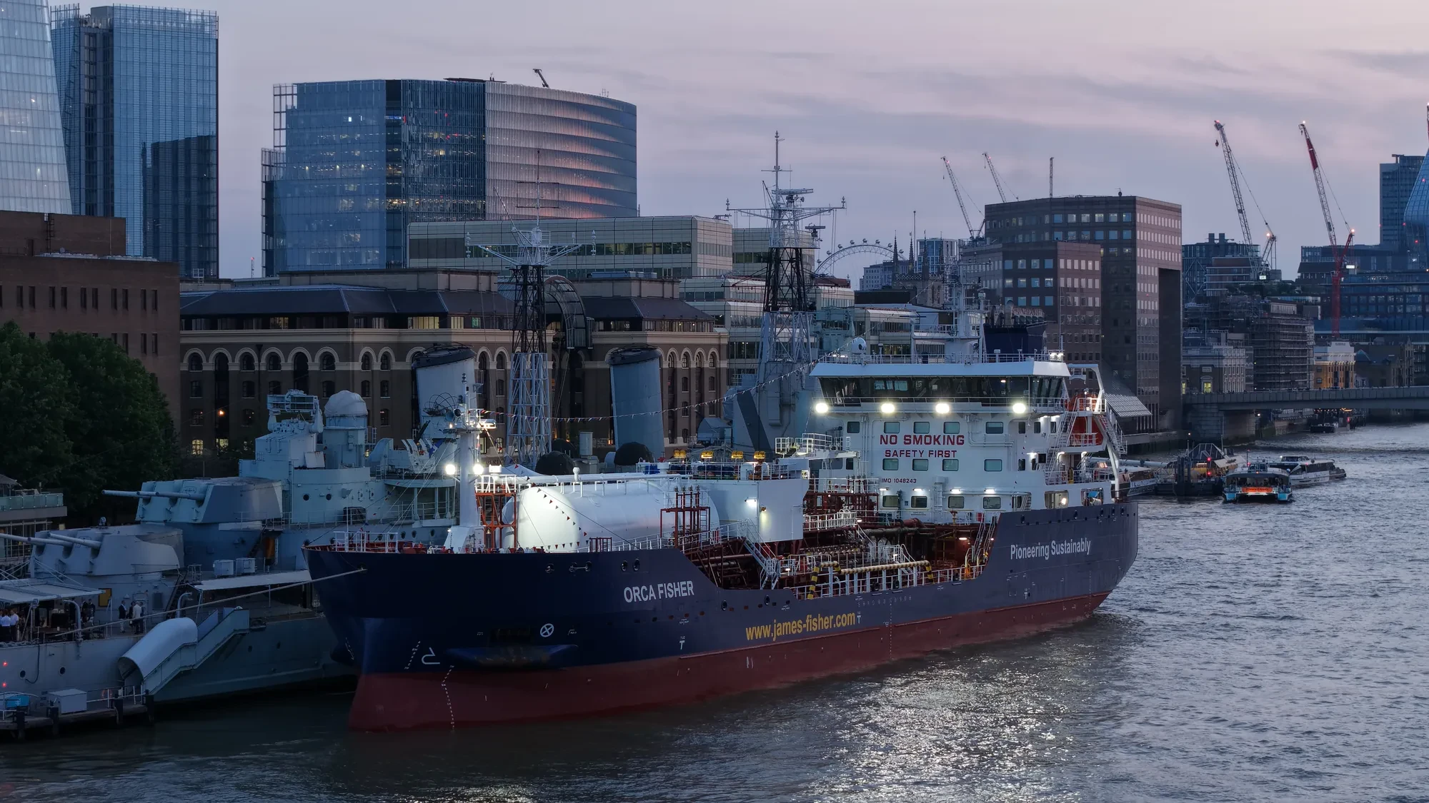 The James Fisher tanker Orca Fisher moored alongside HMS Belfast at dusk with the City of London skyline behind