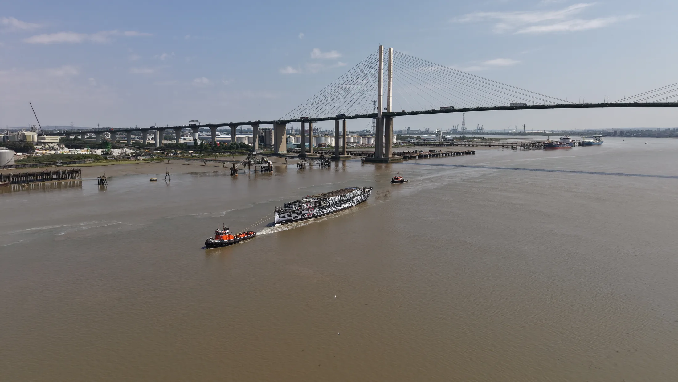 Wide aerial photograph of HMS President under tow on the Thames near the Dartford Crossing, showing the full length of the hull and both attending tugs.