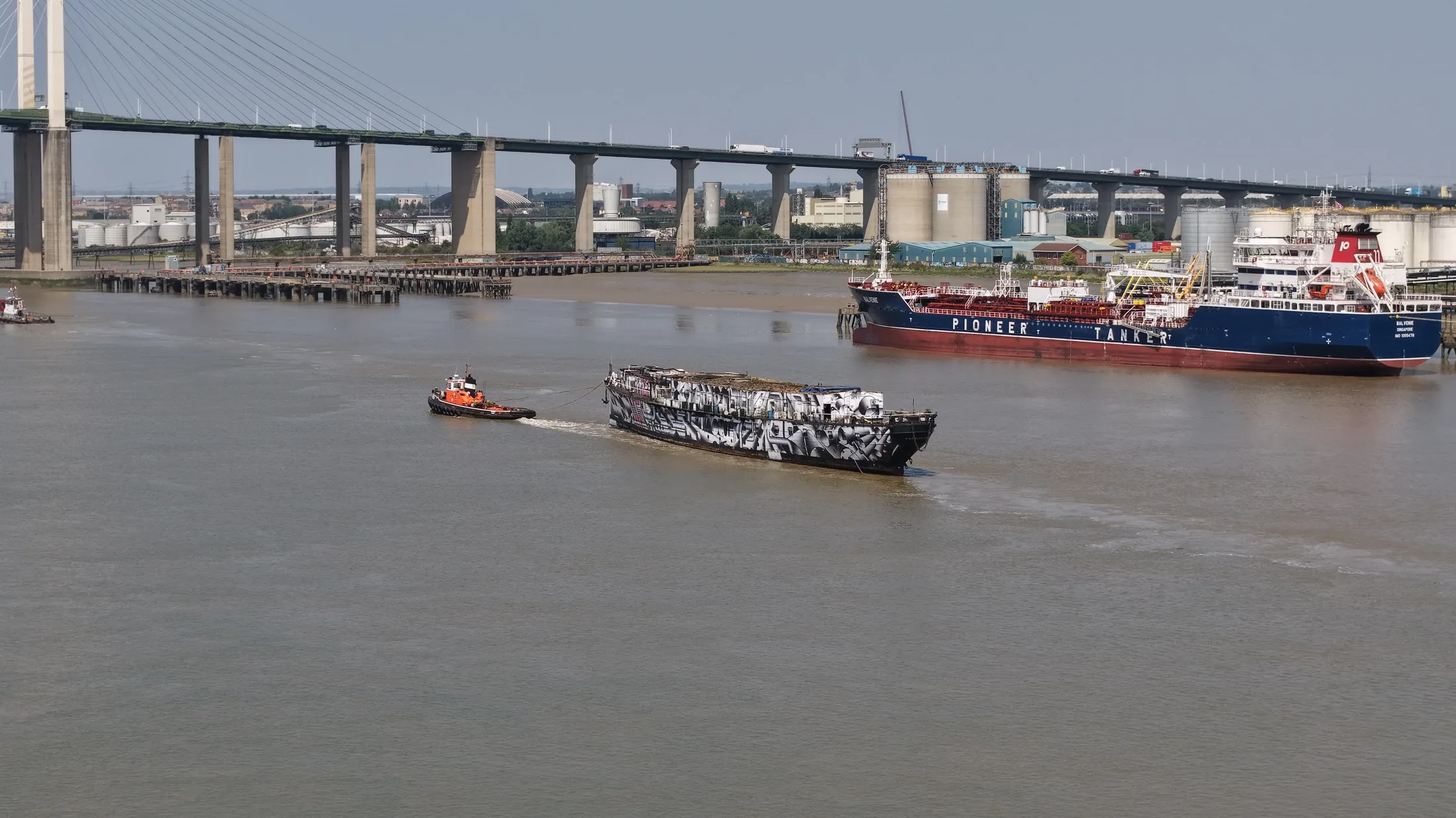 Aerial photograph of HMS President 1918 under tow on the Thames, passing a moored tanker at a riverside jetty near Dartford.