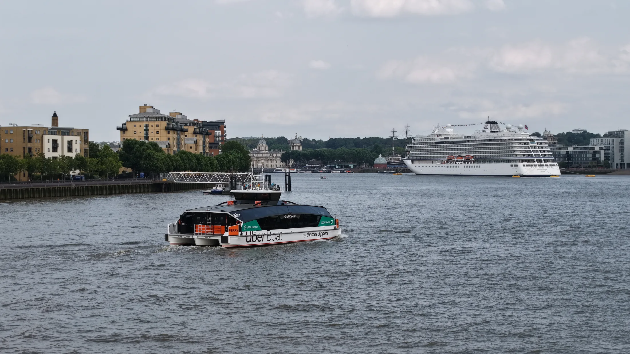 Orbit Clipper under way on the River Thames in east London, with a moored cruise ship visible in the distance downriver.