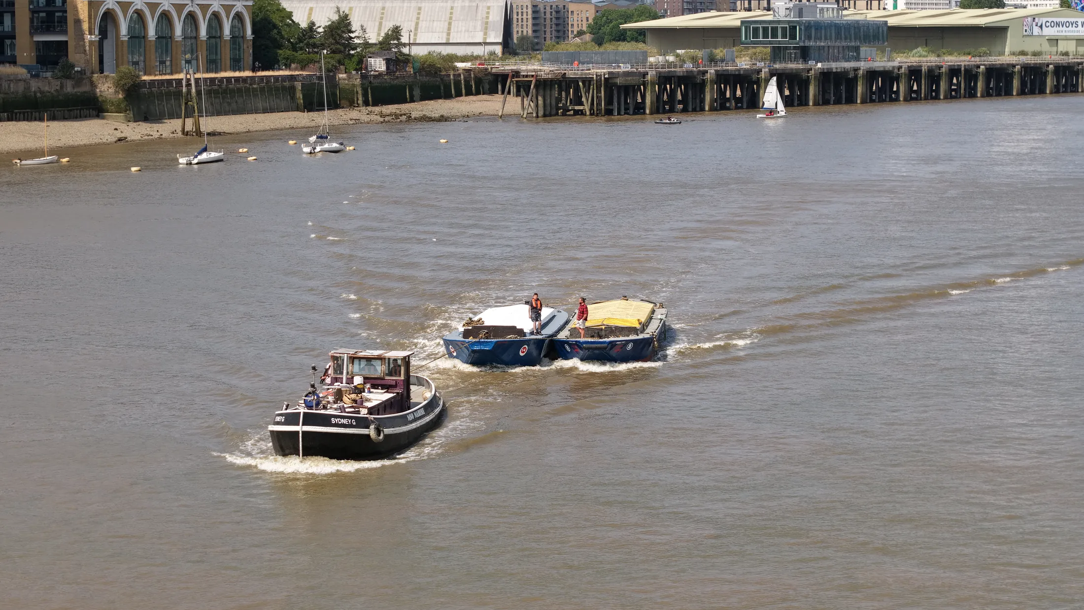 Aerial photograph of a tug towing two racing barges through Greenwich Reach with the north bank in the background.