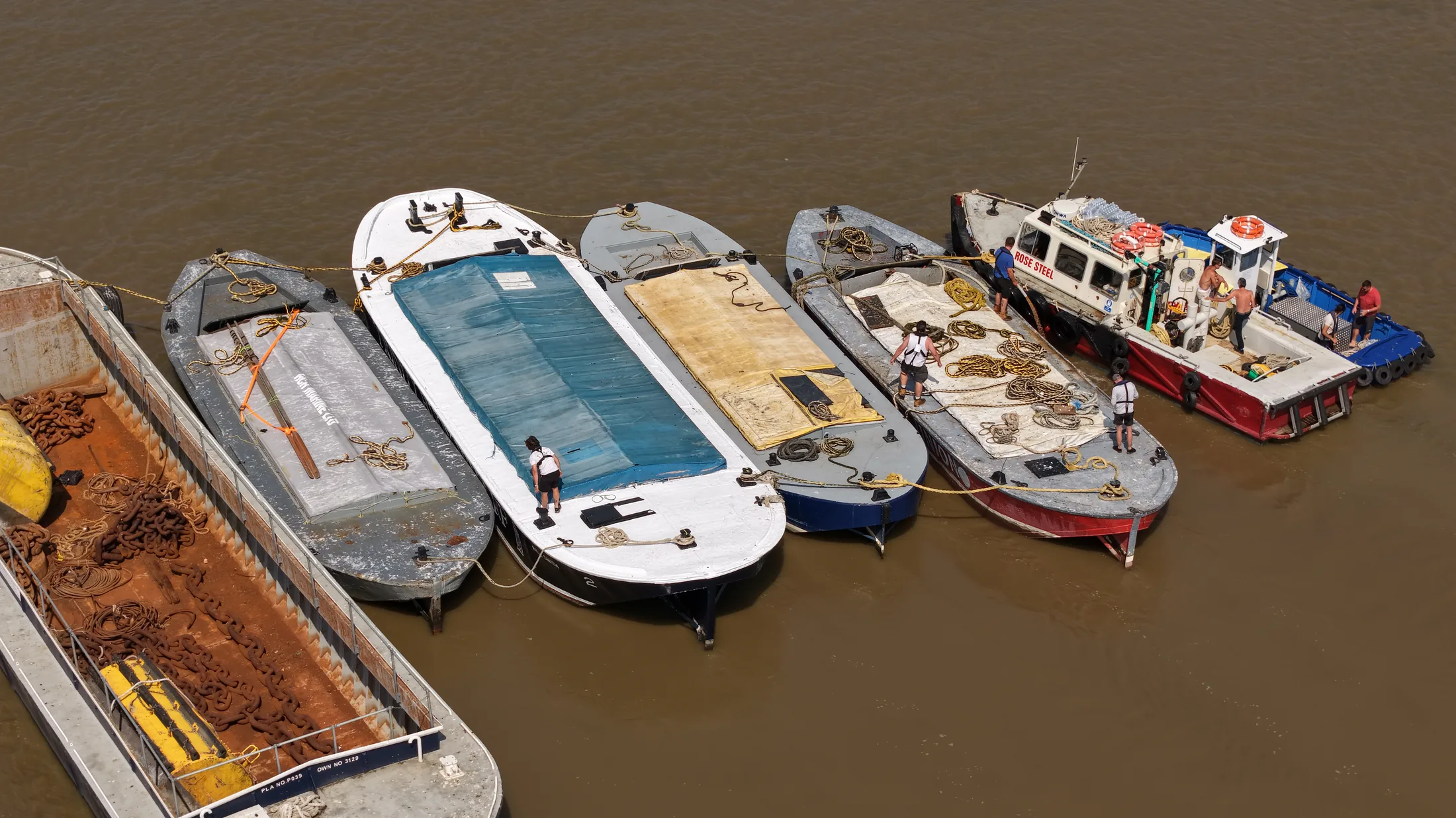 Aerial photograph looking down on Thames barges rafted alongside the tug Rose Steel, with crew members coiling ropes on deck.
