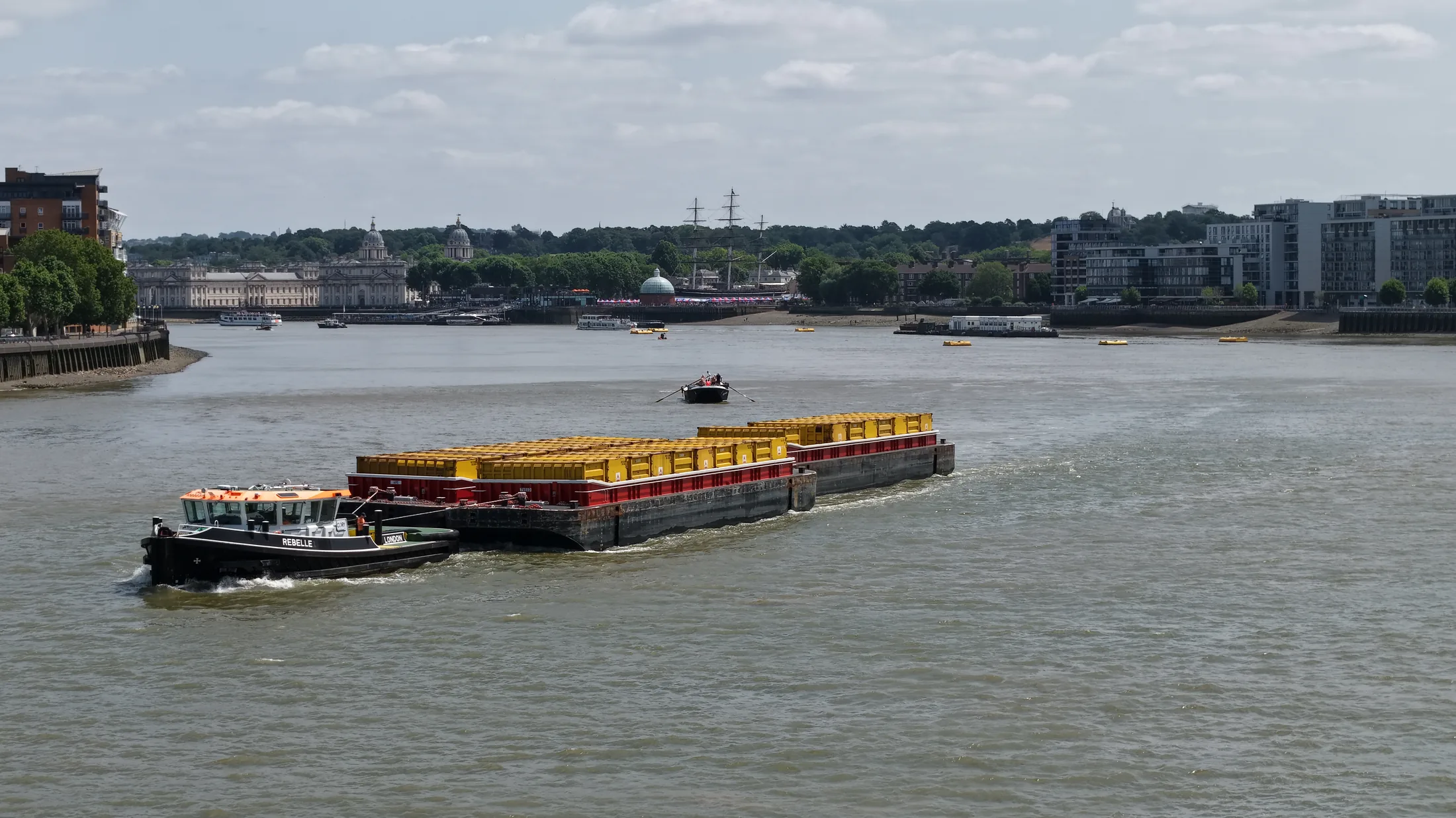 The Cory tug Rebelle towing loaded waste containers on the River Thames during the 2026 barge driving race, with a racing barge under oars in the background.