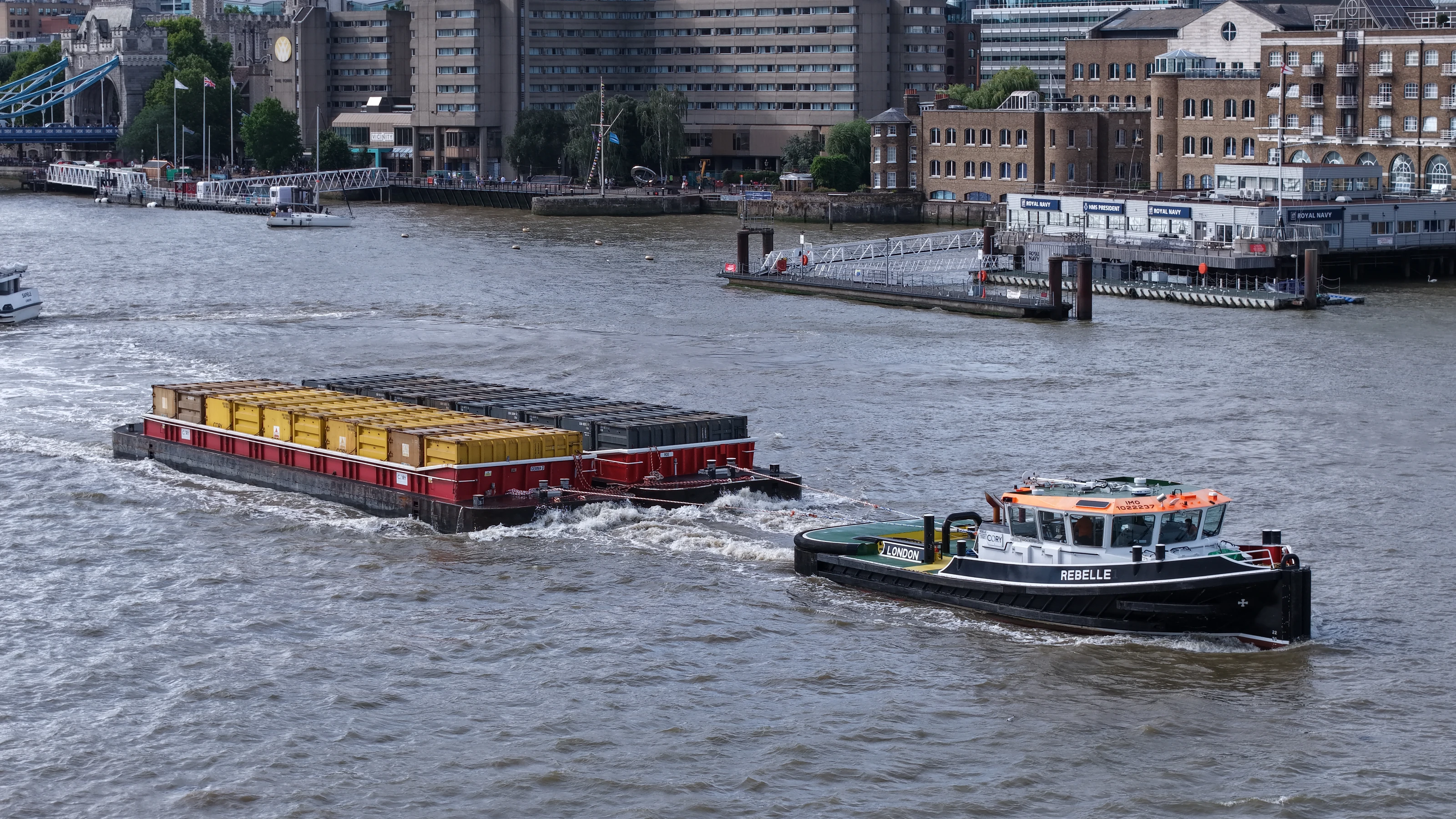 Aerial view of the tug Rebelle towing loaded containers down the tidal Thames past the Bermondsey riverside