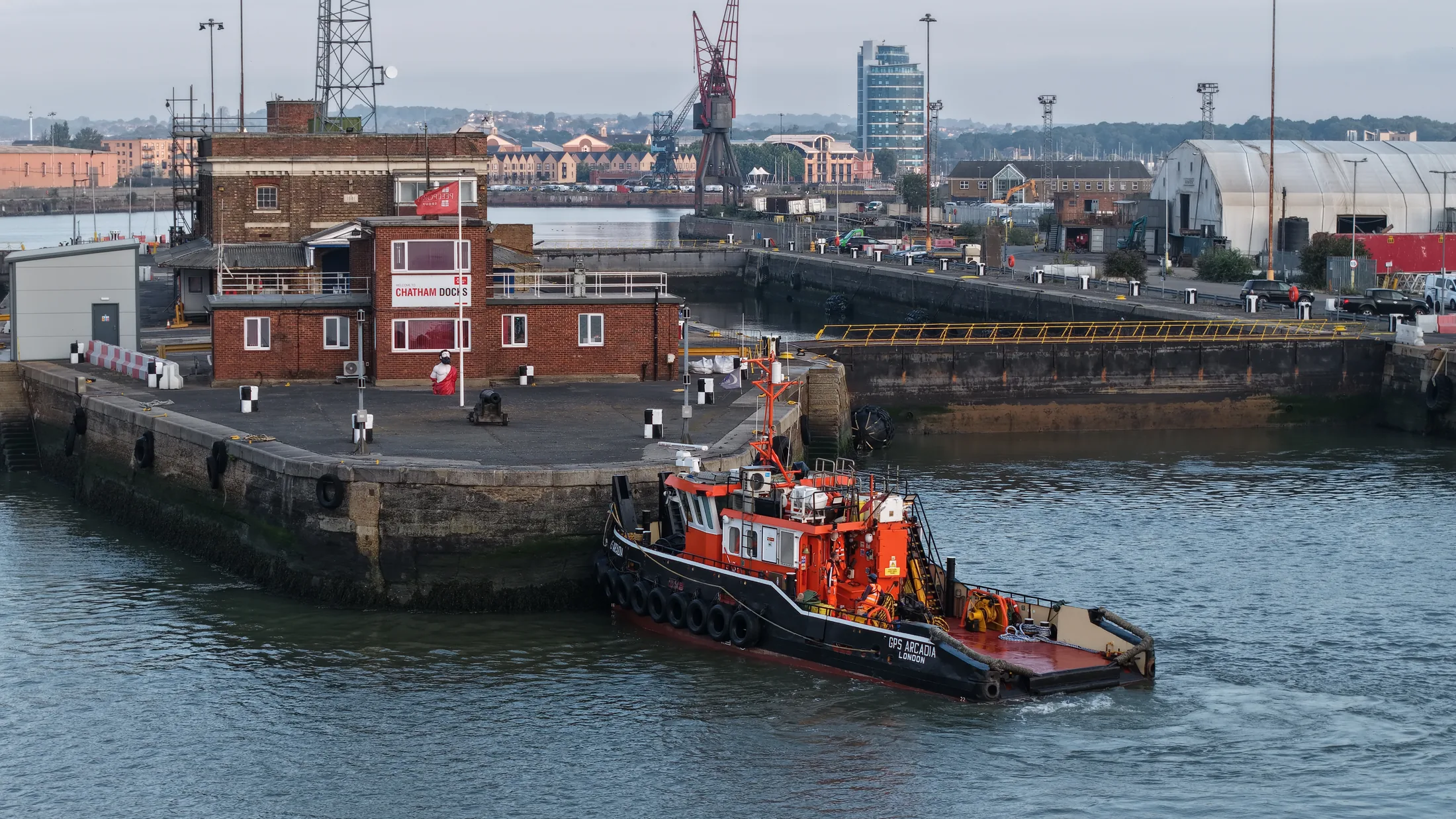 Aerial view of a tug pushing on to the dockyard wall at Chatham, with the historic dockyard buildings and cranes behind.
