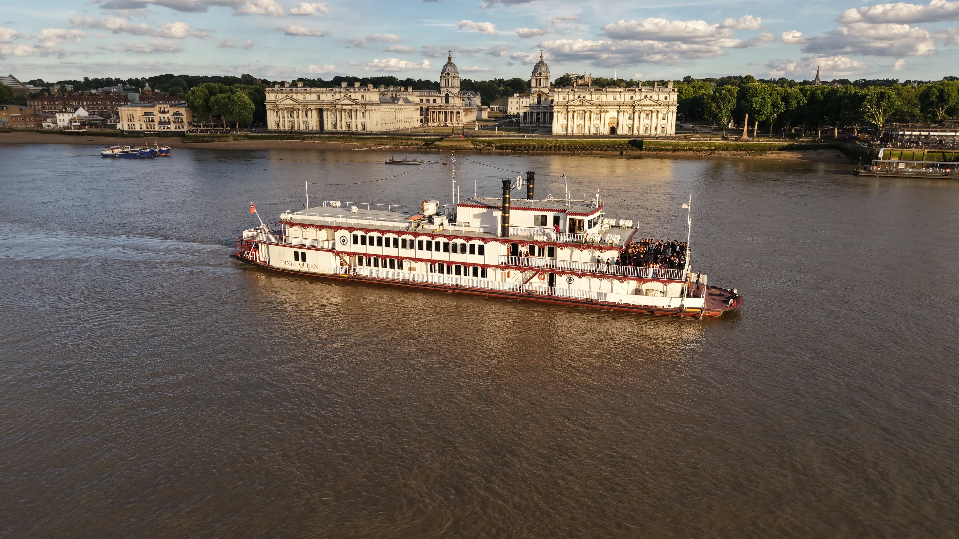 The paddle steamer Dixie Queen passing the Old Royal Naval College at Greenwich in evening light, with guests on the stern deck.