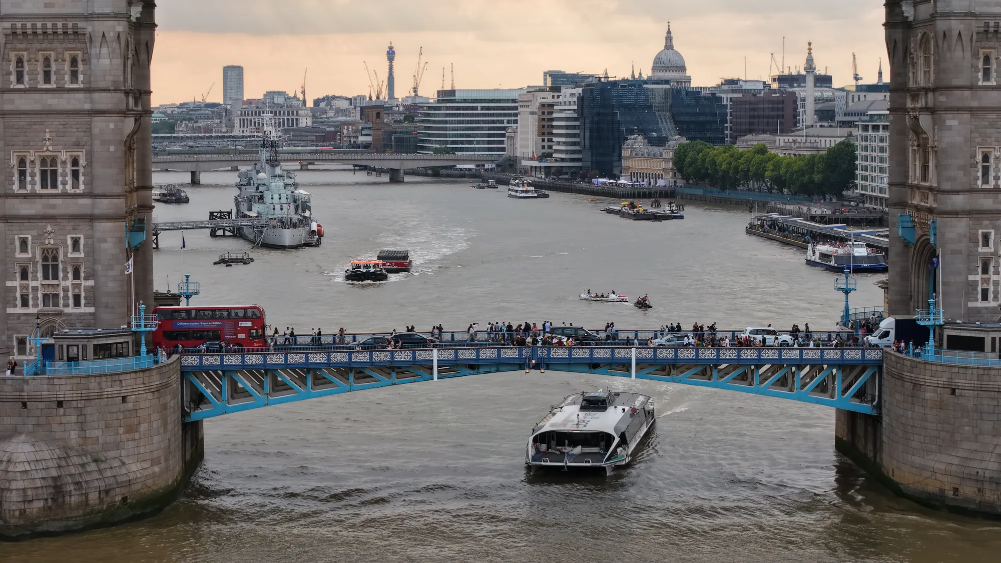 A red London bus crossing Tower Bridge with HMS Belfast and the City skyline beyond, aerial view.