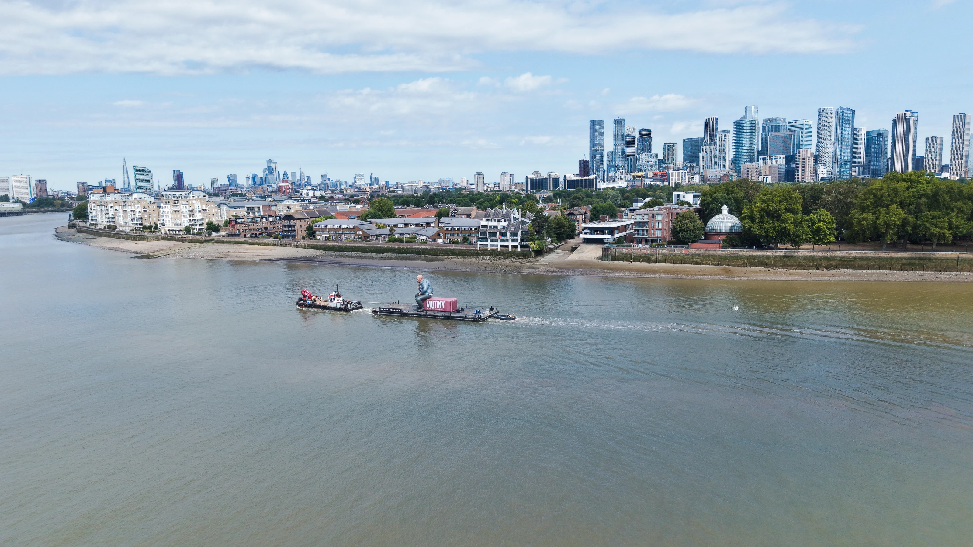 Wide aerial photograph of the Mutiny activation tow in Greenwich Reach, with the Isle of Dogs and the City of London skyline behind