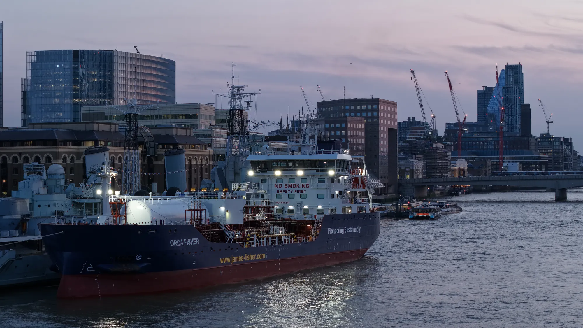 Orca Fisher lit at twilight beside HMS Belfast on the River Thames, City of London in the background