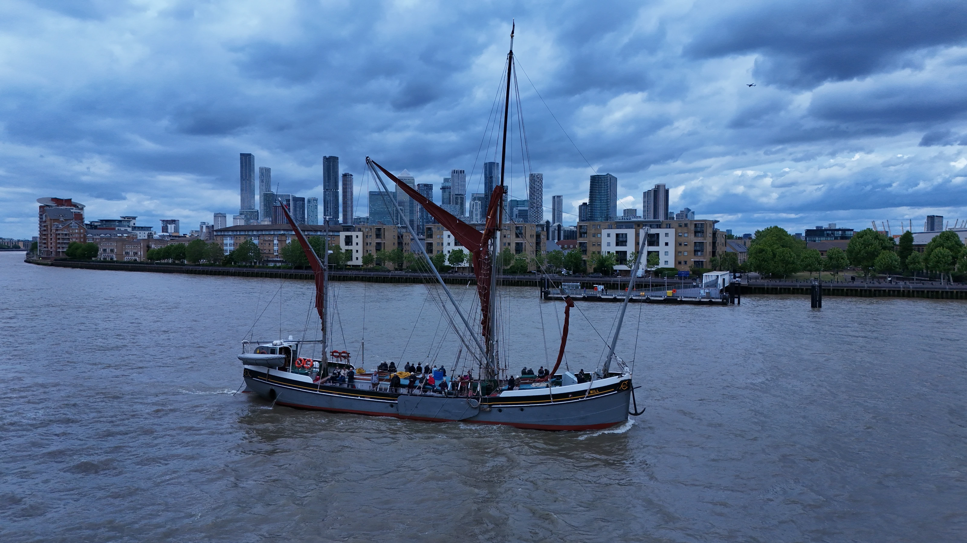 Aerial view of the sailing barge Will on the River Thames in London, tan sails brailed and passengers aboard.